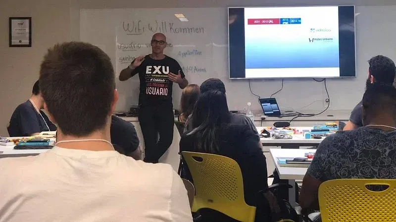 Horácio Soares apresentando para grupo em sala de treinamento, com whiteboard e tela projetando interface de aplicativo, participantes sentados em cadeiras amarelas.