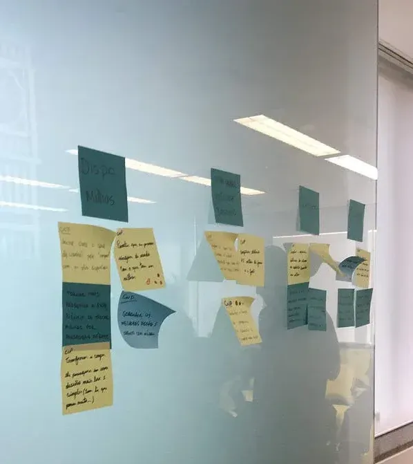 Parede de vidro com post-its amarelos e azuis organizados em colunas durante sessão de design thinking ou workshop colaborativo em escritório.