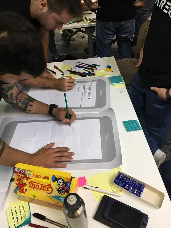 Pessoa com tatuagens desenhando wireframes de dispositivos móveis em papel durante workshop colaborativo, com materiais de design espalhados na mesa incluindo marcadores, post-its e protótipos